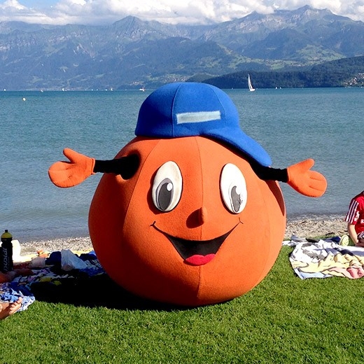 Badi-Tour 2025 - Ein oranges, grosses Conci-Plüschtier steht in einer Badi auf der Wiese am Wasser.