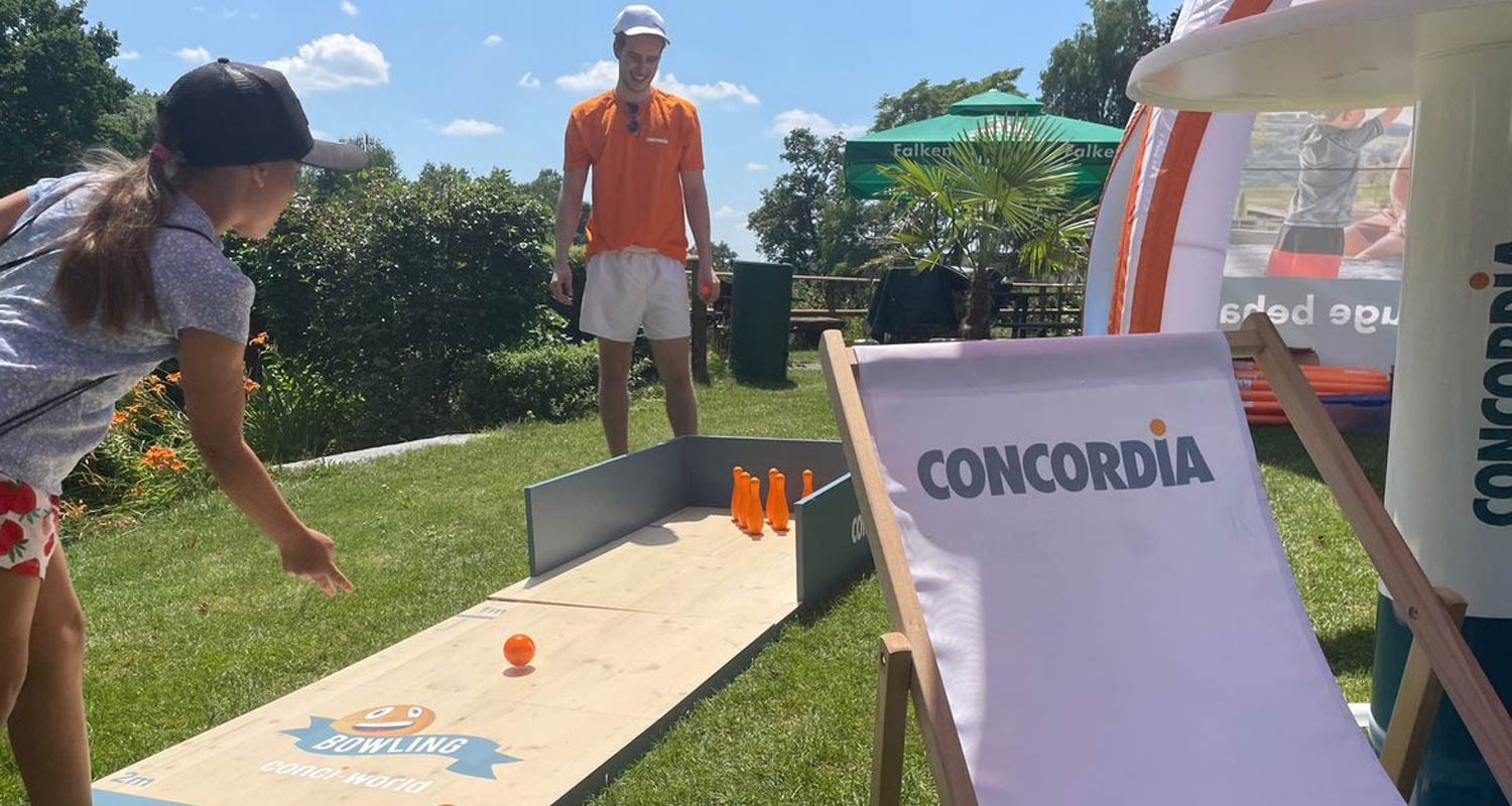 Badi-Tour 2025 - Ein Mädchen spielt am CONCORDIA-Stand der Badi-Tour Mini-Bowling, ein Helfer der CONCORDIA betreut sie.