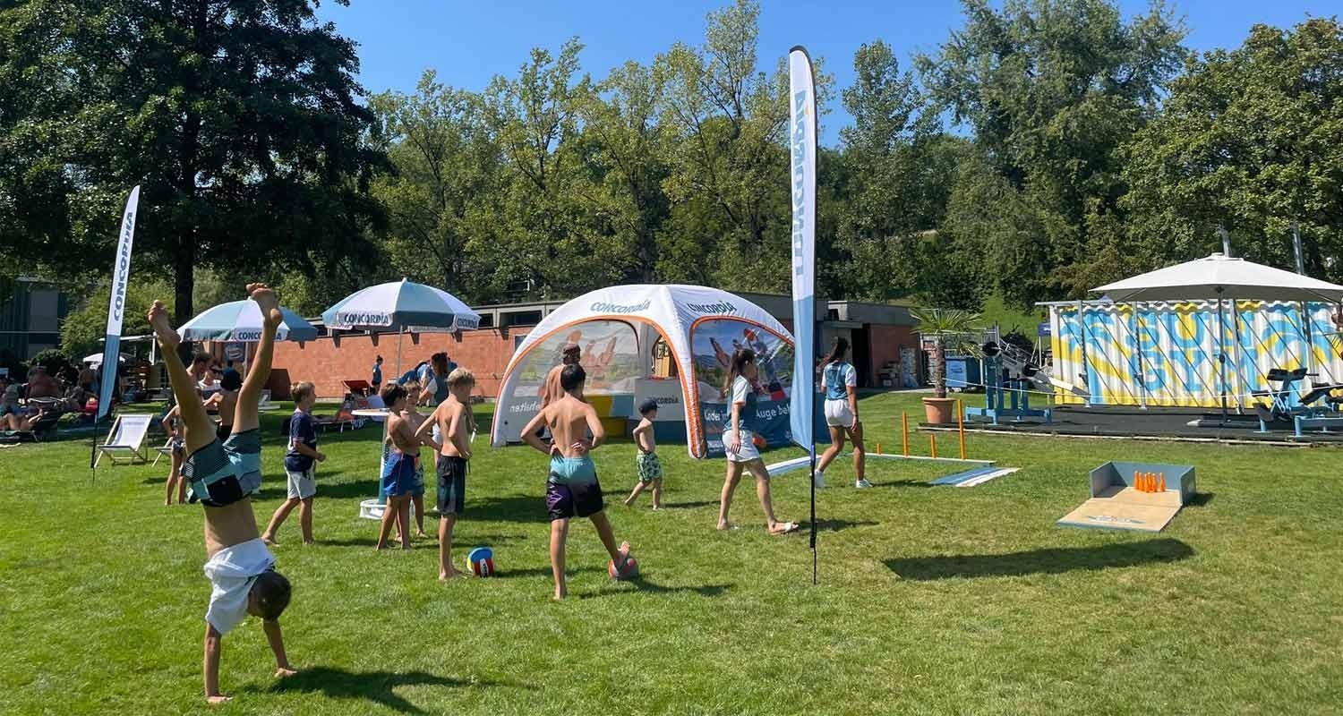 Badi-Tour 2025 - Eine Gruppe Kinder spielt auf einer Wiese vor dem Badi-Tour Stand der CONCORDIA.