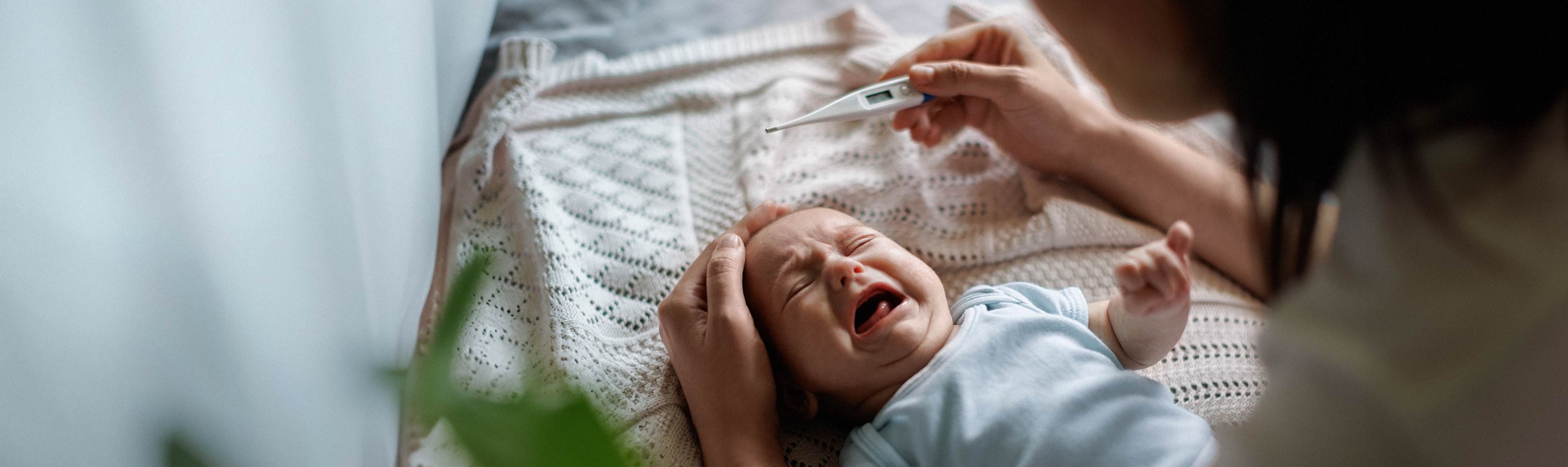 Mein Kind ist krank - Baby weint und liegt im Bett, die Mutter hält einen Fiebermesser