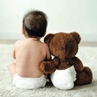 Baby sitzt neben Teddybär. Der Teddybär umarmt das Baby herzlich