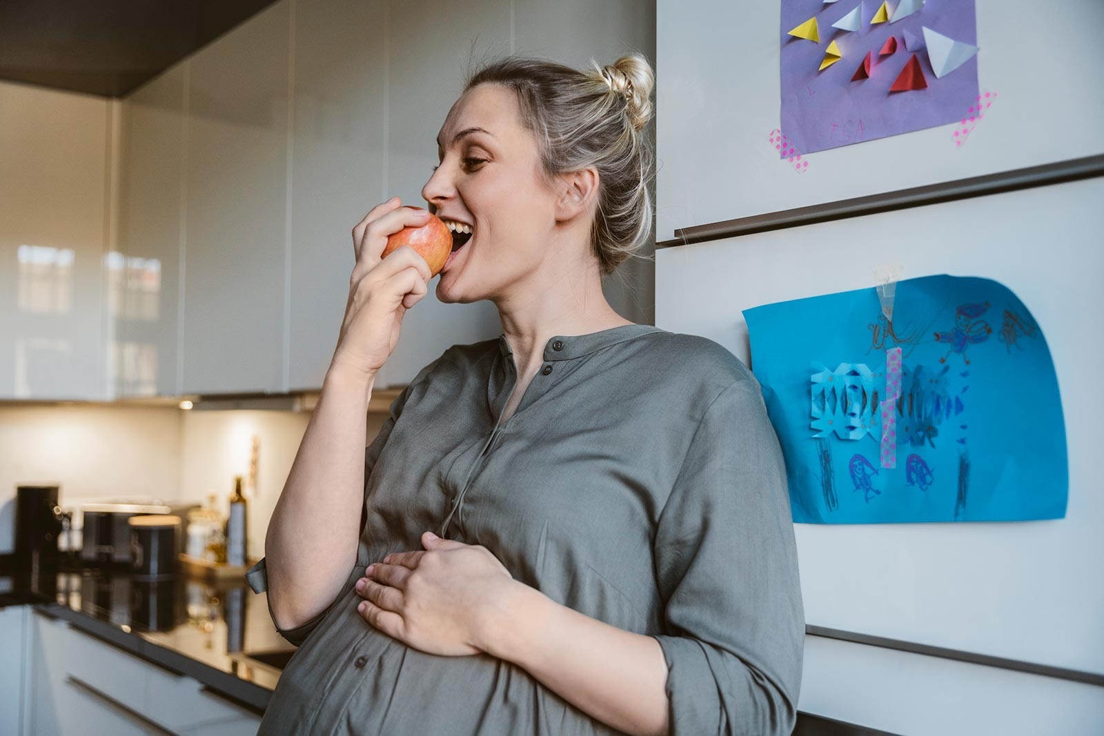 Gesunde Ernährung Schwangerschaft – Schwangere Frau steht in der Küche und beisst in einen Apfel