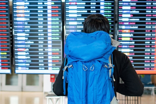 Backpacker startet seine Weltreise. Er trägt einen blauen Rucksack und steht vor einem Fluginformationsplan an einer Abflugtafel.