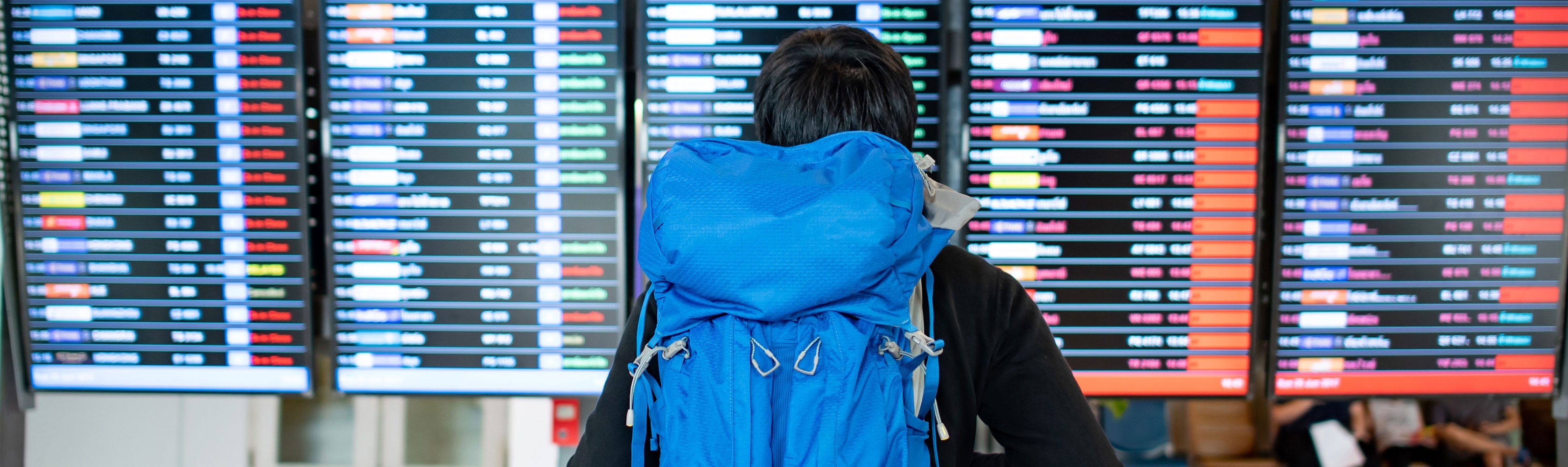 Backpacker startet seine Weltreise. Er trägt einen blauen Rucksack und steht vor einem Fluginformationsplan an einer Abflugstafel.