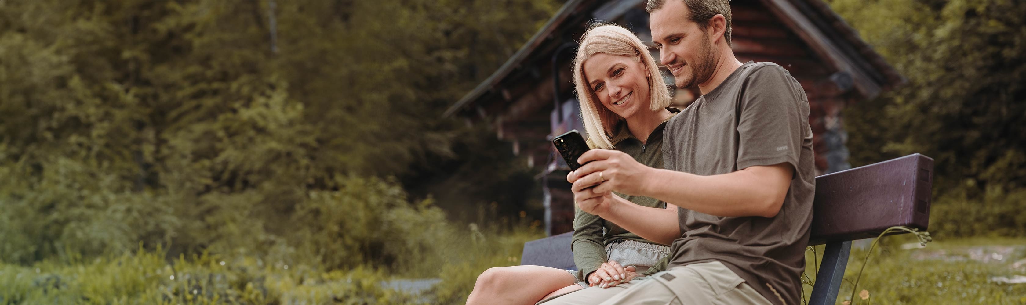 myCONCORDIA Registrierung Kundenportal und App - Frau sitzt mit Mann am See und zeigt ihm das Kundenportal auf dem Smartphone.