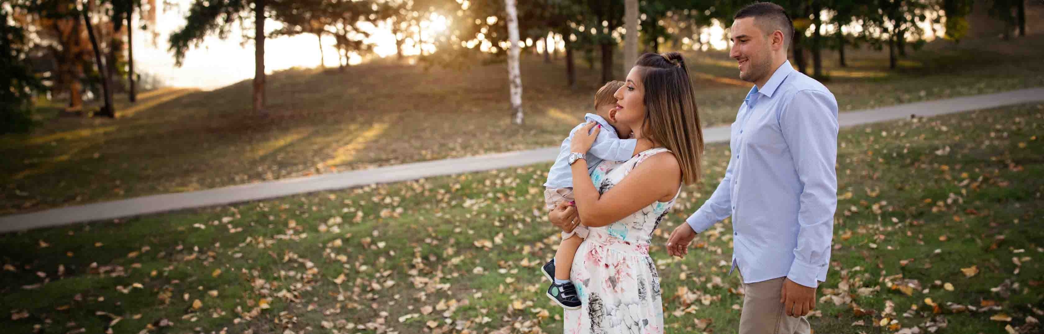 Familje shqiptare me 3 ne park. Nëna mban fëmijën në krahë.