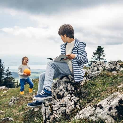 Knabe sitzt auf einem Stein mit einem Buch. Seine Schwester spielt im Hintergrund mit einem orangen Ball.