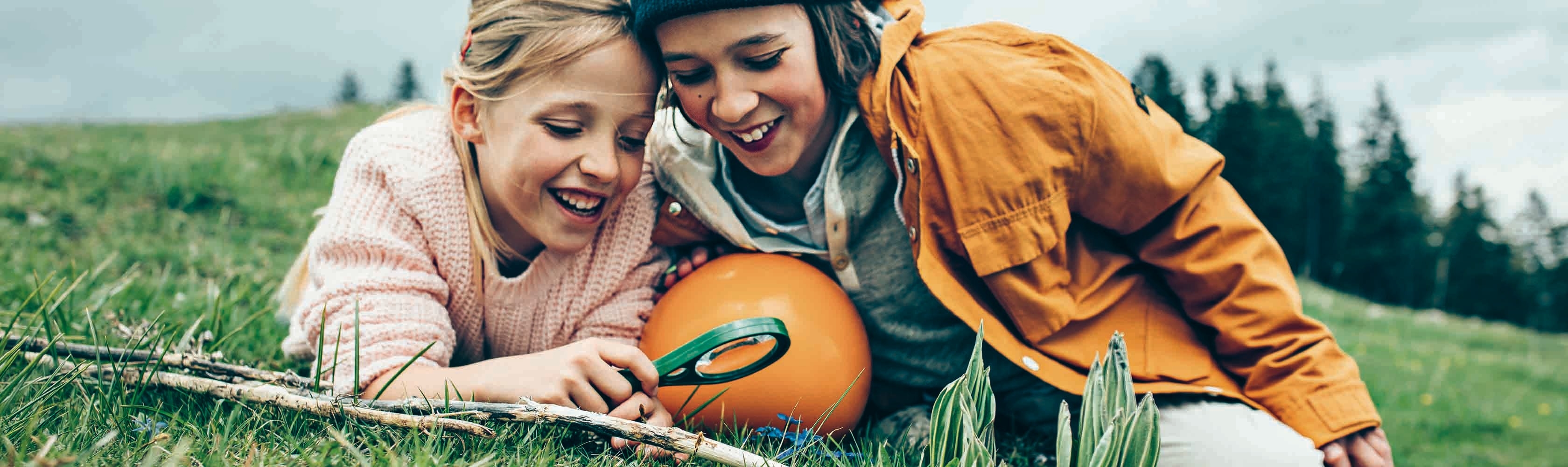 2 Kinder schauen mit einer Lupe in die Wiese, sie knien auf dem Boden und halten den orangen Concordia Ball.