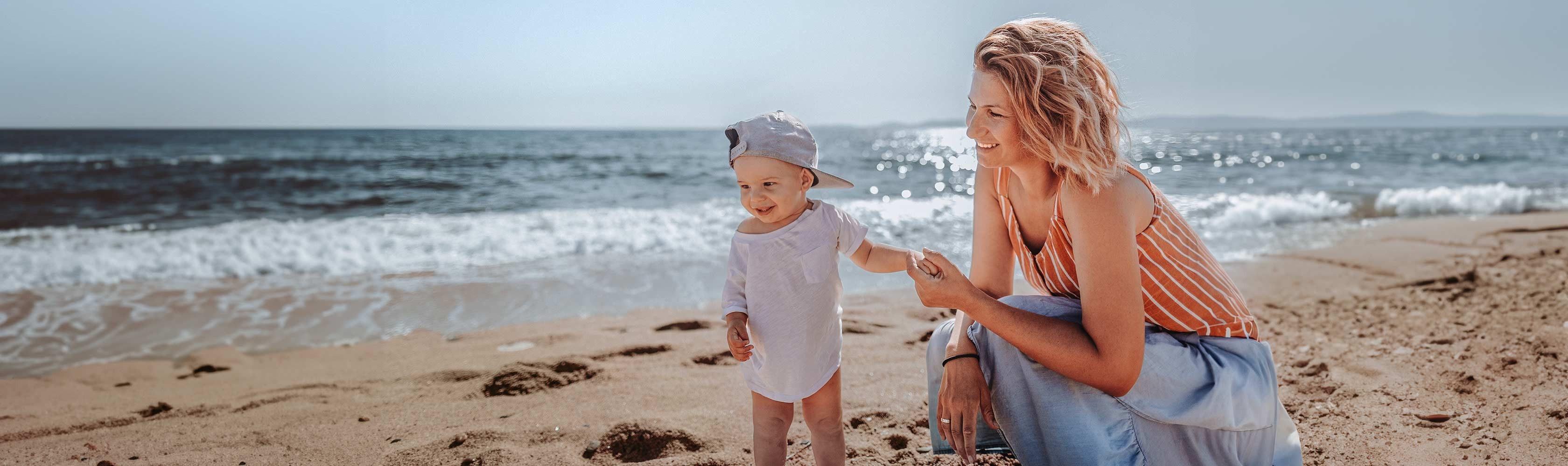 Sonnenstich - Mutter steht mit ihrem Kleinkind mit Sonnenhut am Strand, im Hintergrund das Meer