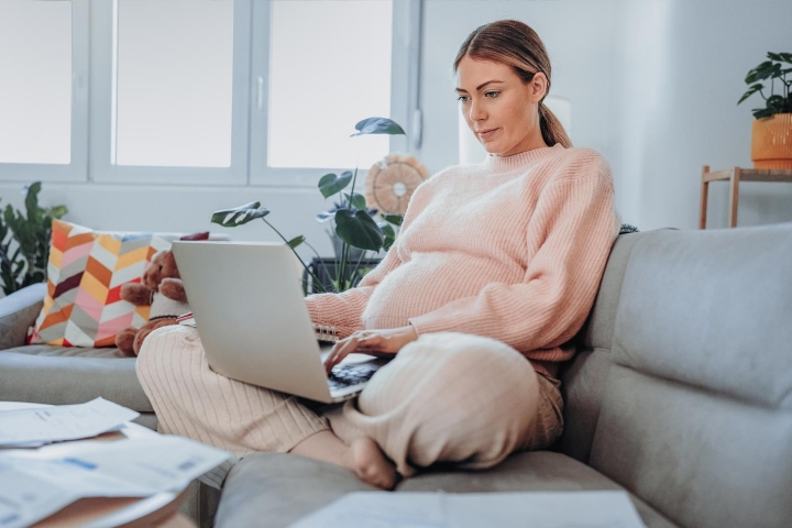 A pregnant woman sits on the sofa with her laptop and paperwork