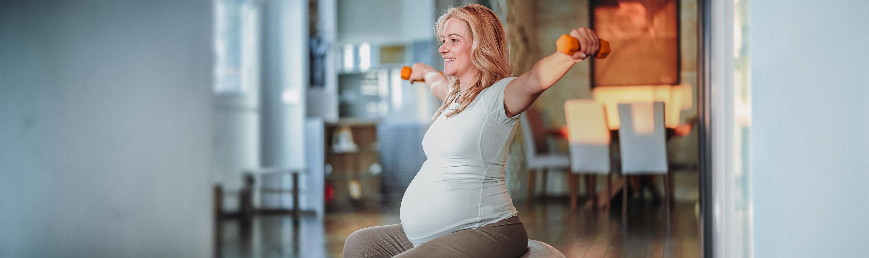 Exercise and pregnancy – a woman sits on an exercise ball and stretches her arms out sideways with a weight in each hand