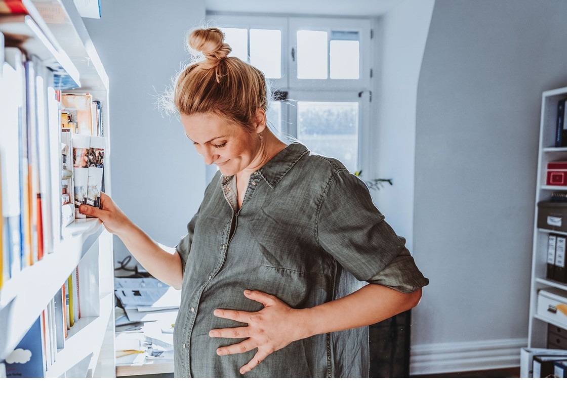Birth preparation– a pregnant woman stands next to a bookshelf with one hand on her tummy