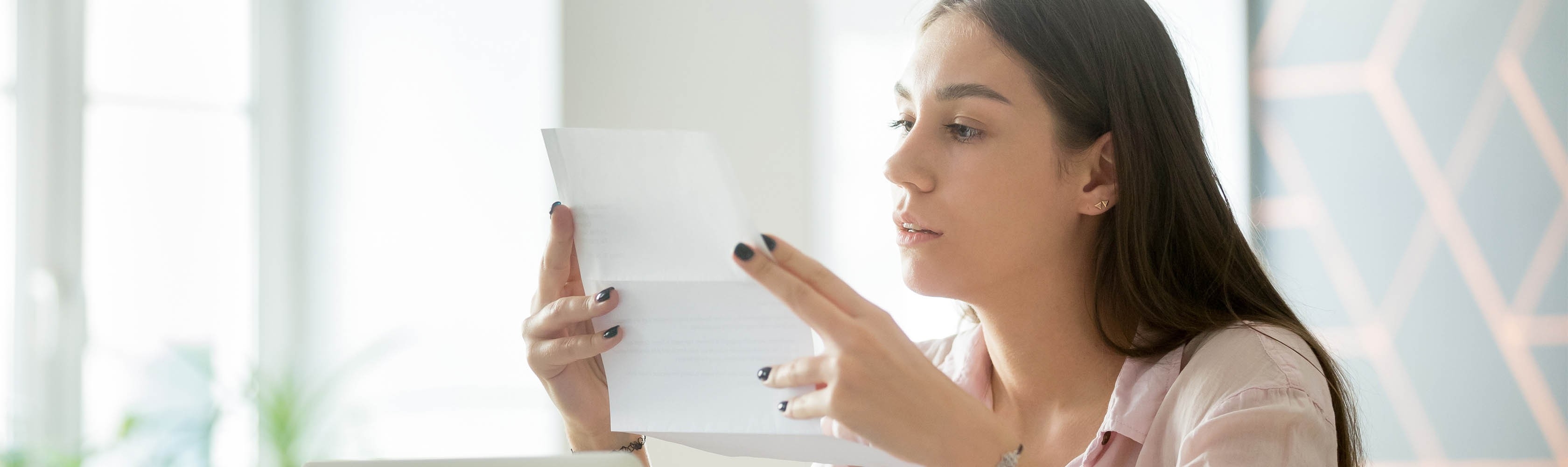 Assise à son bureau, une jeune femme tient à la main un courrier de sa caisse-maladie l’informant des changements survenant à la majorité.