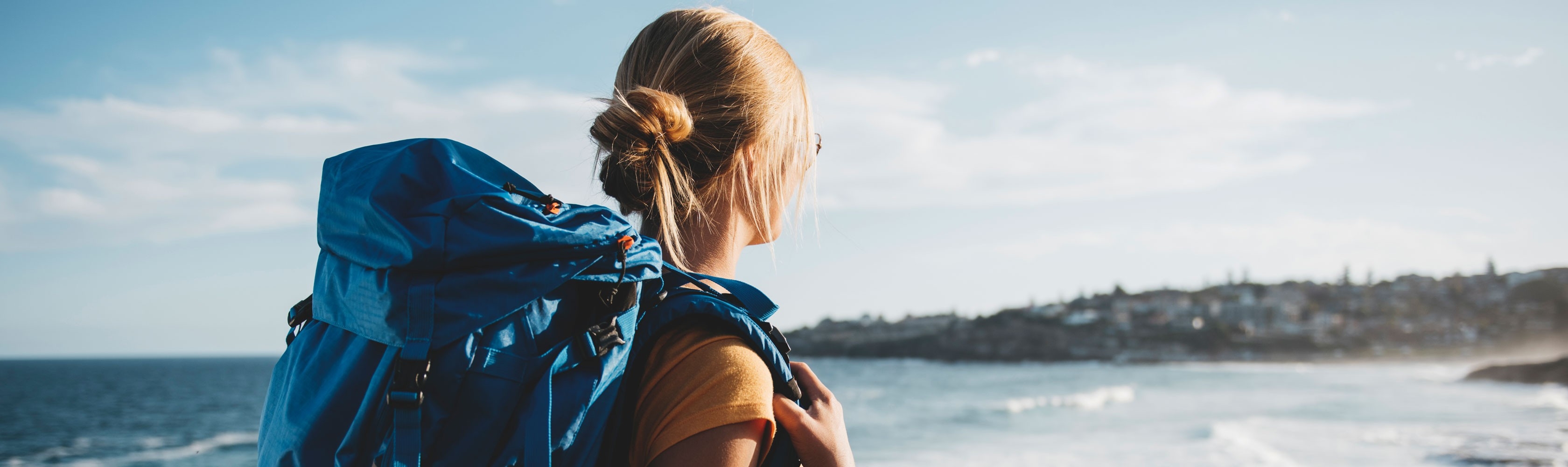 Femme en voyage autour du monde. Elle porte un sac à dos bleu et regarde la mer.