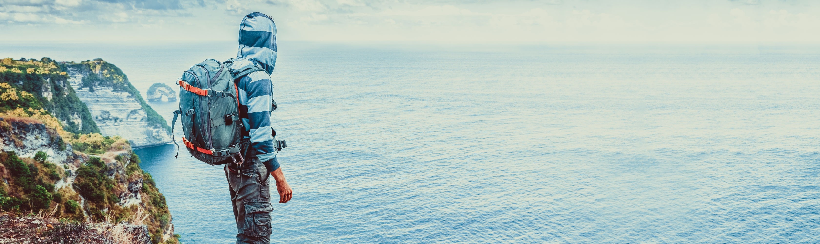 Un homme en voyage autour du monde : il se tient au bord d'une falaise à Bali, porte un sac à dos et regarde la mer.
