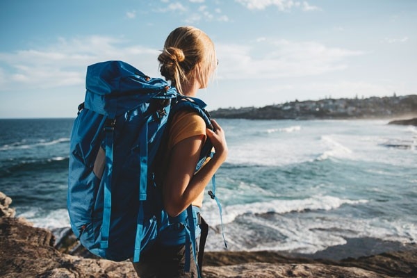 Femme en voyage autour du monde. Elle porte un sac à dos bleu et regarde la mer.