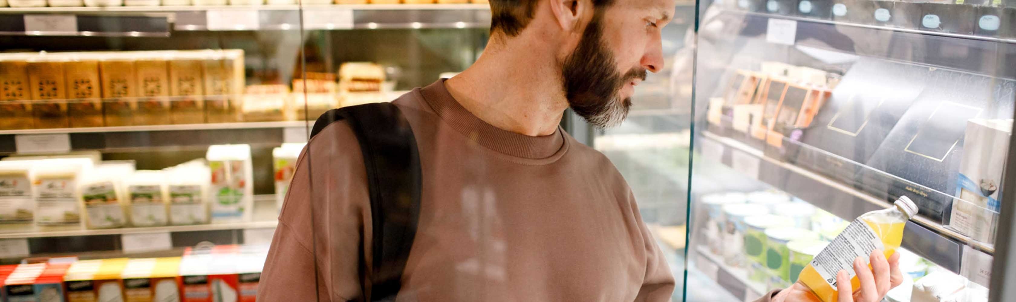 Un homme se tient devant le rayon frais du supermarché et lit la liste des ingrédients sur une bouteille de jus d'orange.