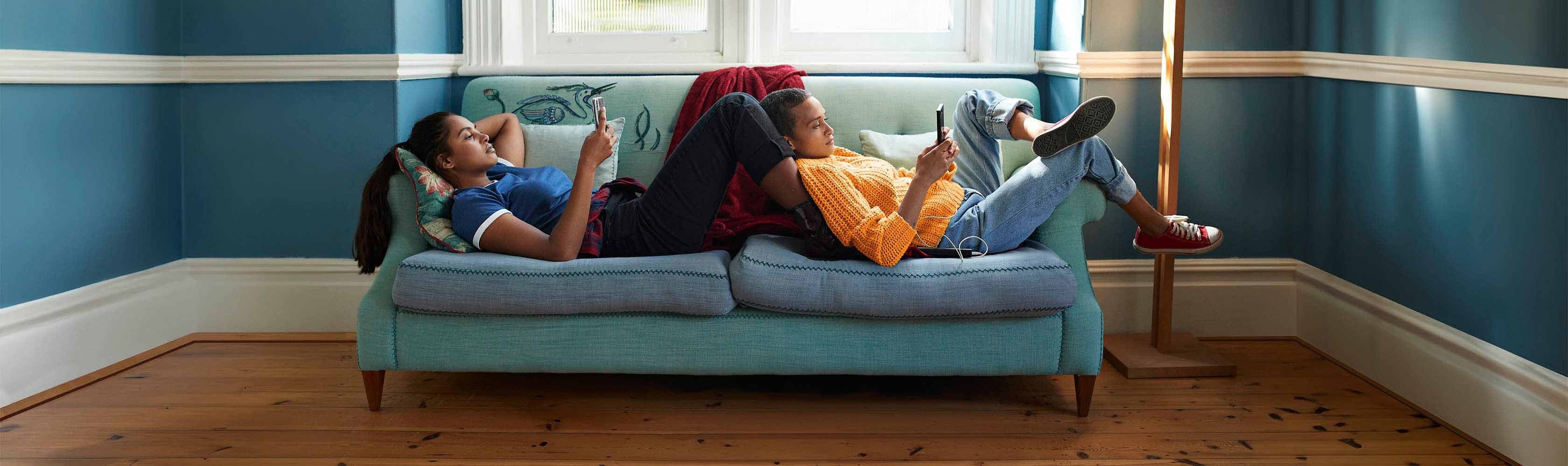 Rivista online - Due donne guardano il cellulare mentre sono sdraiate su un piccolo divano blu.