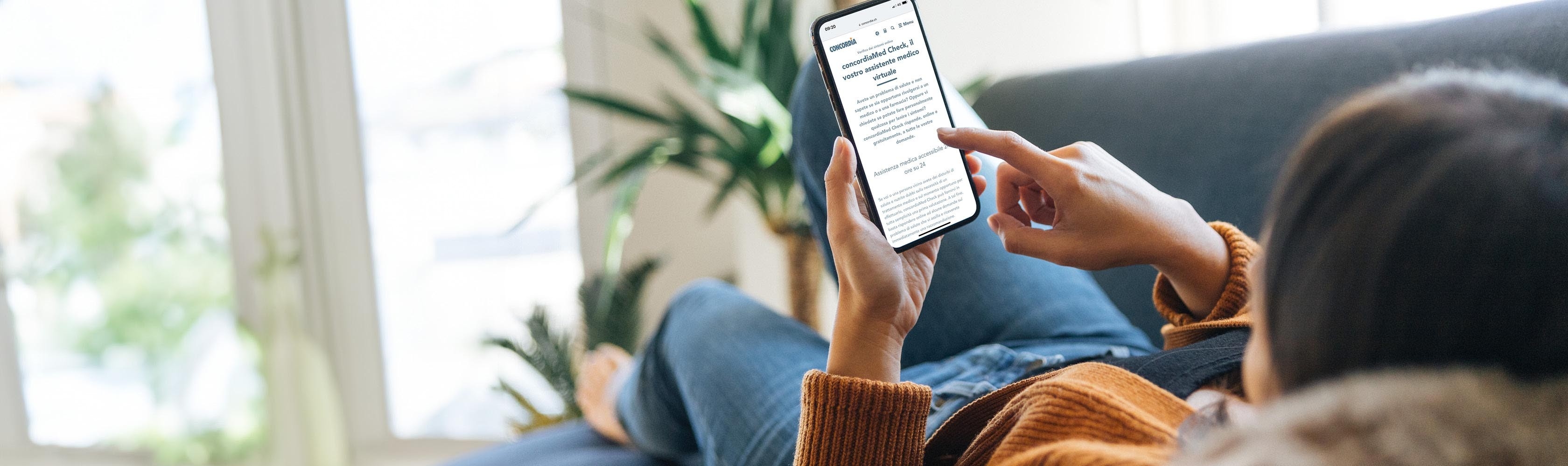 Una giovane donna legge sul cellulare le informazioni su concordiaMed Check, sdraiata sul divano.