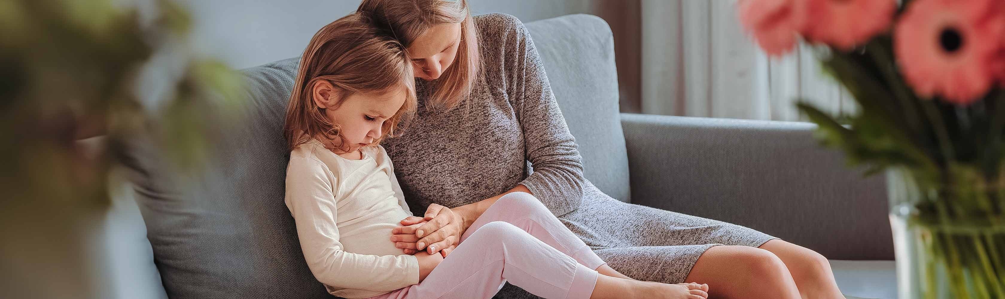 Bauchweh - Kind sitzt mit der Mutter auf dem Sofa und hält sich den Bauch
