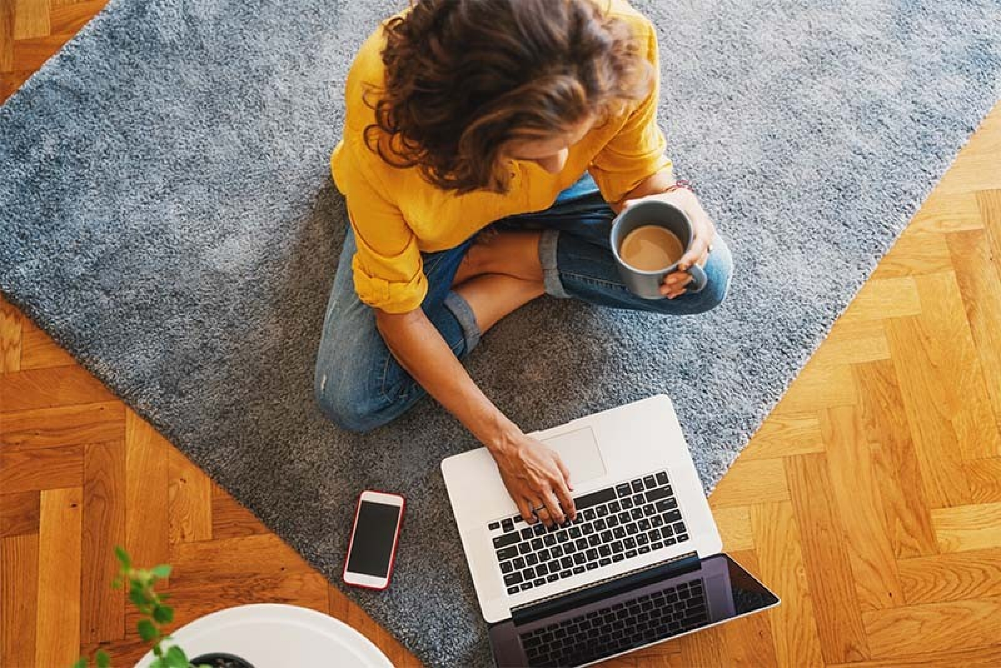 Una donna è seduta nel suo salotto, con una tazza di caffè in mano, e sta lavorando al computer portatile.