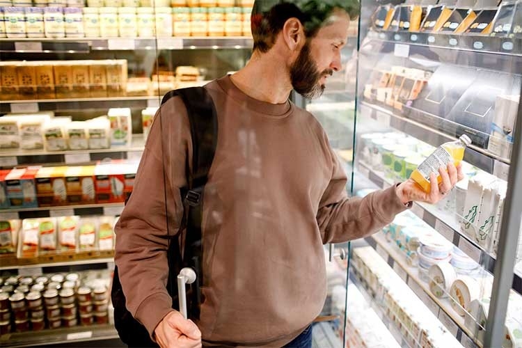 Un uomo è davanti allo scaffale dei prodotti refrigerati al supermercato e sta leggendo gli ingredienti riportati su una bottiglia di succo d'arancia.