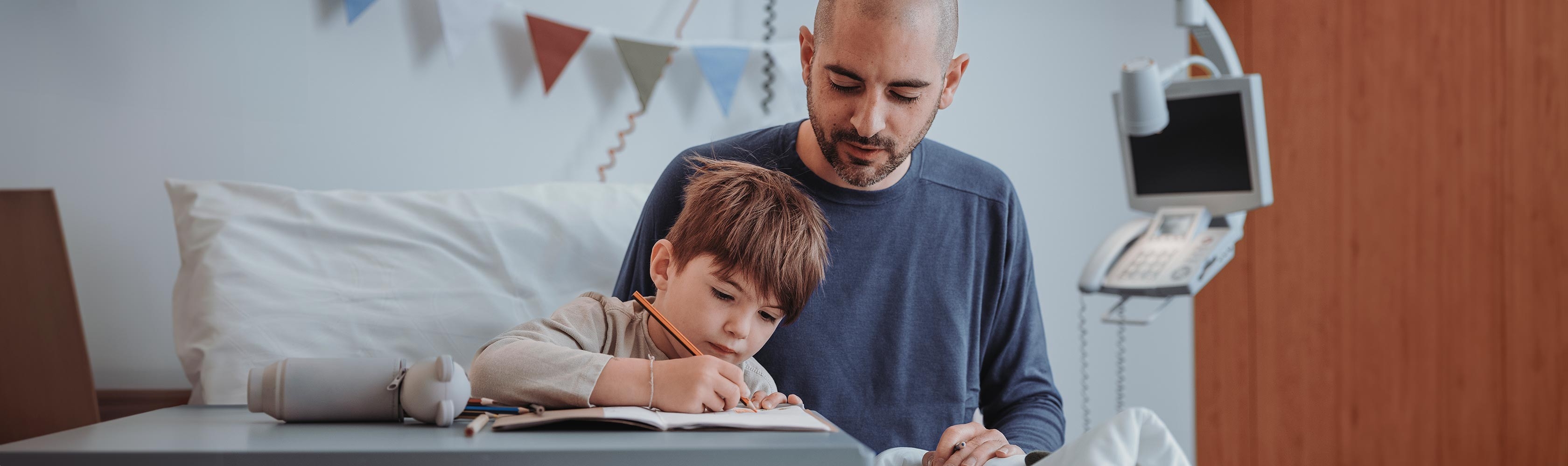 Knabe sitzt auf einem Stein und liest ein Buch. Im Hintergrund steht ein Mädchen mit einem organgen Ball.