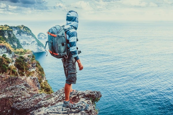 Un uomo in viaggio intorno al mondo: è in piedi sul bordo di una scogliera a Bali, porta uno zaino e guarda il mare.
