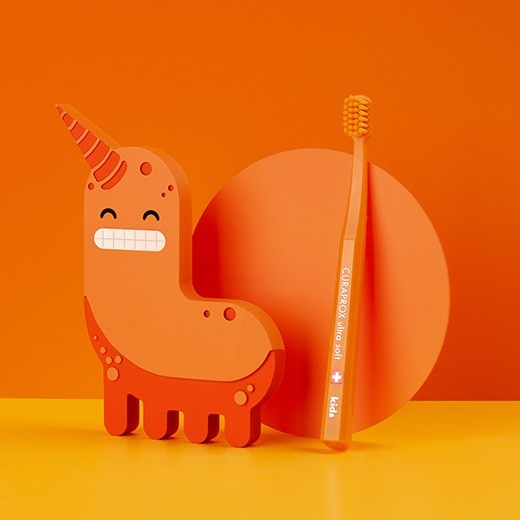 Bestellung Kinderzahnbürste - Eine orange Kinderzahnbürste von Curaprox neben einem orangem Masskottchen
