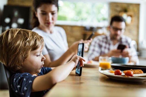 Gesünder leben: Mutter, Vater und Kind sitzen am Esstisch, alle mit dem Handy in der Hand. Eltern haben Vorbildfunktion für ihre Kinder, auch beim Online- und Medienkonsum.