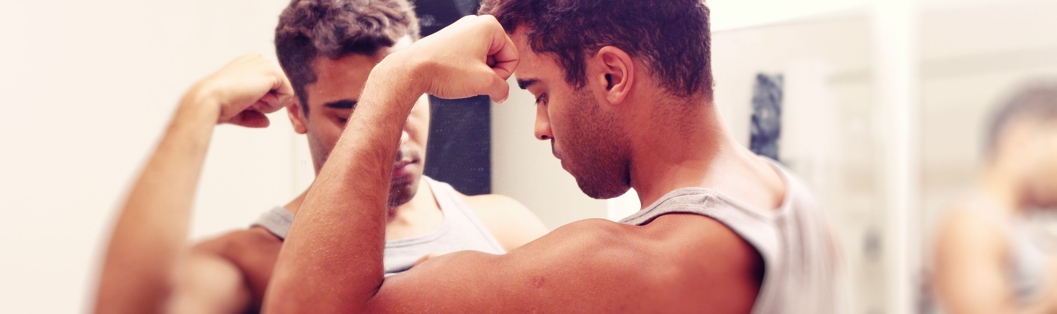 Ein junger Mann betrachtet im Spiegel seine Muskeln am Oberarm.