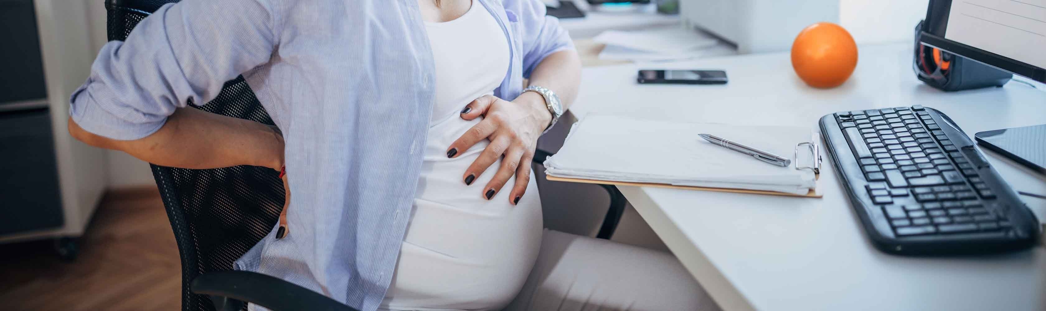 Frau sitzt am Arbeitsplatz und hat Rückenschmerzen in der Schwangerschaft.