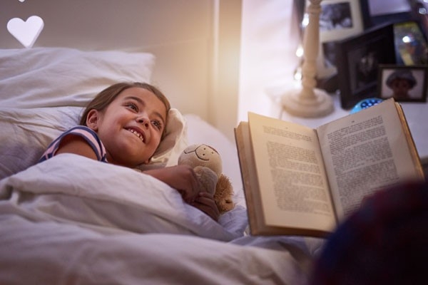 La fille est au lit avec un ours en peluche. Sa mère lit une histoire tirée d'un livre.