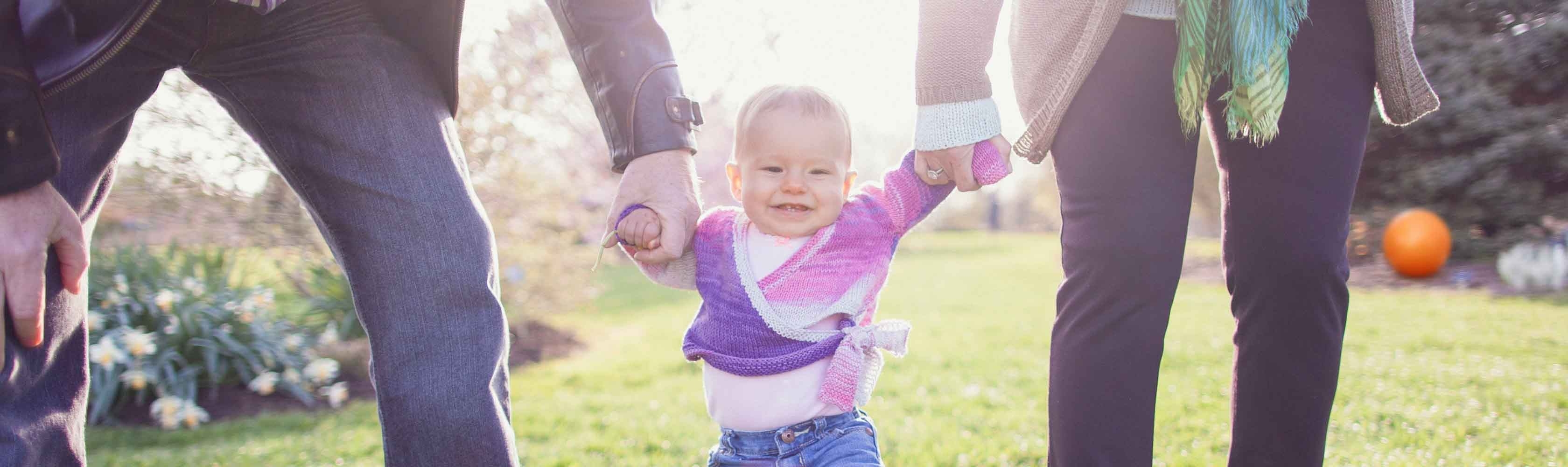 Versicherung während der Schwangerschaft und nach der Geburt – Eltern laufen mit Baby über Wiese.