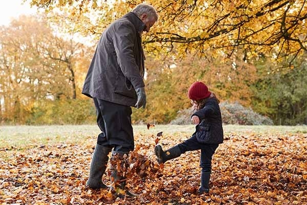 Un bambino gioca con il nonno tra le foglie secche cadute al suolo in un bosco. Negli inverni miti il rischio zecche è reale.